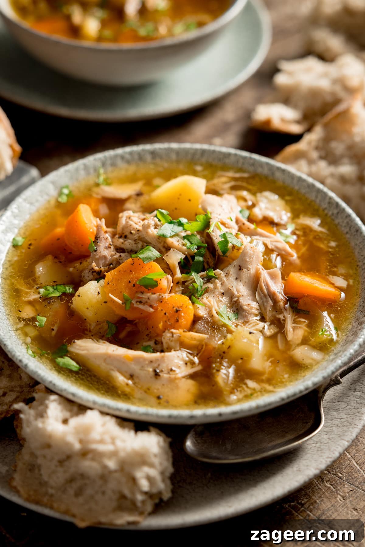 Hearty Slow Cooker Chicken and Veggie Bowl 5 A tall, close-up image of slow cooker chicken and vegetable soup in a light bowl, placed on a wooden table. A piece of bread and a spoon accompany the bowl. In the soft background, another bowl of soup and more bread are visible, highlighting a comforting meal setting.