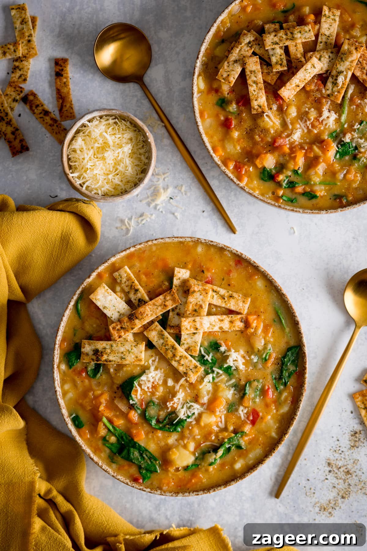 Hearty Slow Cooker Lentil Bowl 5 A tall, appetizing image featuring two bowls of creamy red lentil soup, each generously topped with crunchy crispy tortilla strips, presented on a light background. A golden spoon rests in one bowl, and around them are scattered extra tortilla strips and a small bowl of grated cheese, along with a cheerful yellow napkin, creating a cozy and inviting scene.
