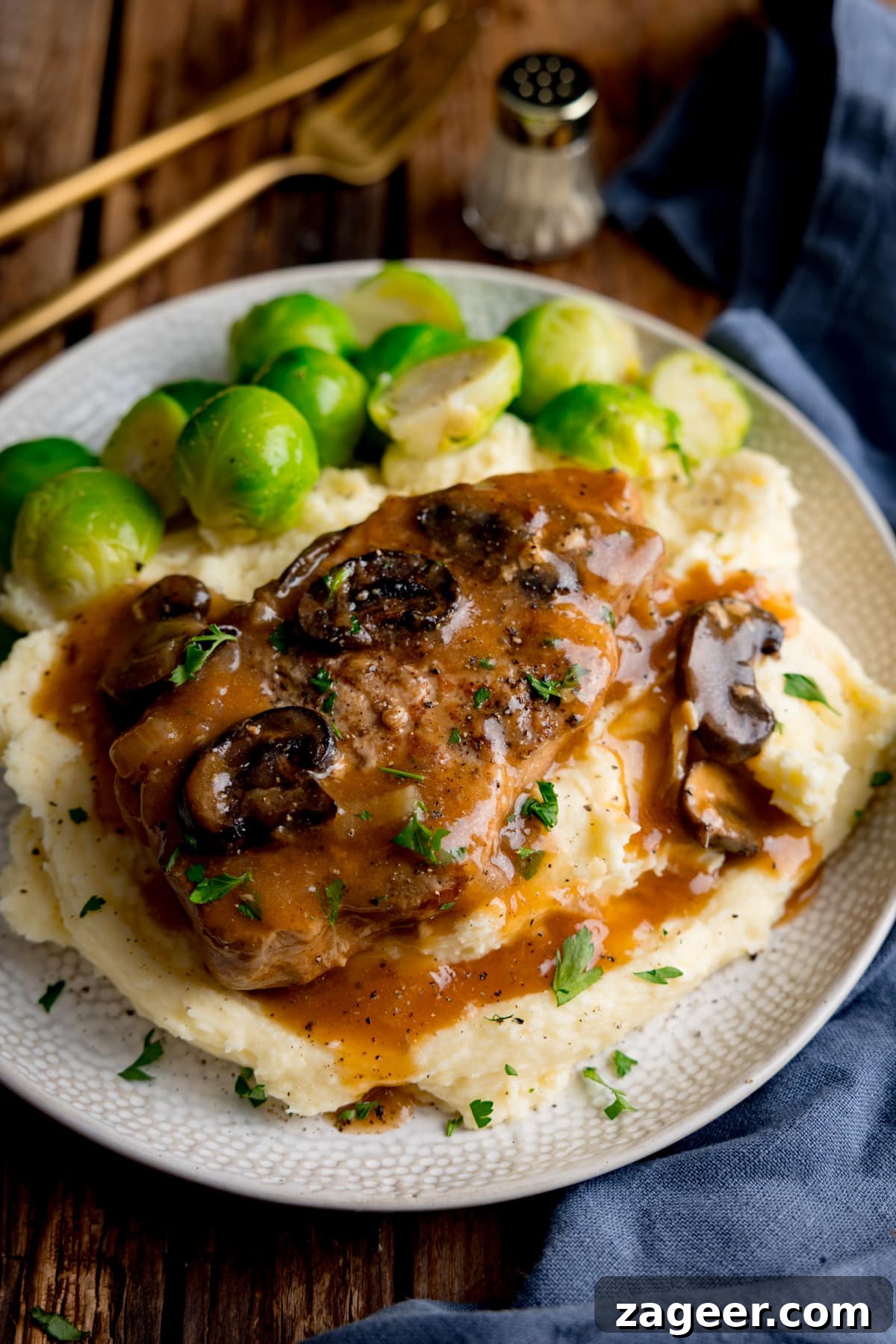 Fall-Apart Slow Cooker Smothered Pork Chops 2 Slow cooker smothered pork chop with gravy and mushrooms on top of a pile of mashed potato, next to sprouts on a white plate. The plate is on a wooden background next to a blue napkin. There is gold cutlery and a small salt shaker at the top of the image.