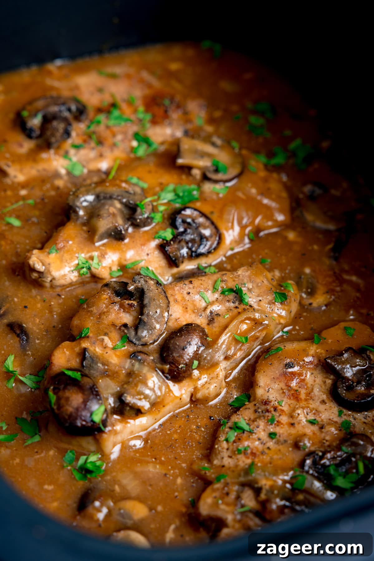 Fall-Apart Slow Cooker Smothered Pork Chops 4 Tall close-up image of smothered pork chops with mushrooms and gravy in the pan of a slow cooker. There is chopped parsley sprinkled on top.