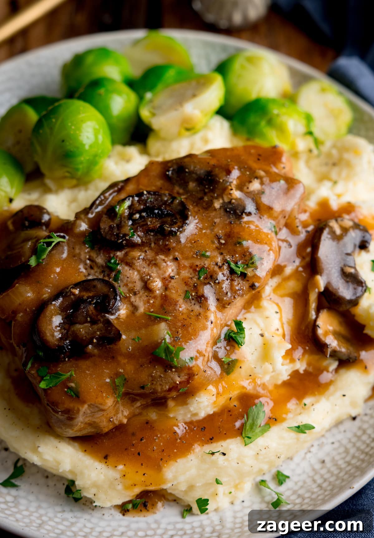 Fall-Apart Slow Cooker Smothered Pork Chops 5 Slow cooker smothered pork chop with gravy and mushrooms on top of a pile of mashed potato, next to sprouts on a white plate. The plate is on a wooden background next to a blue napkin. There is some gold cutlery at the top of the image.