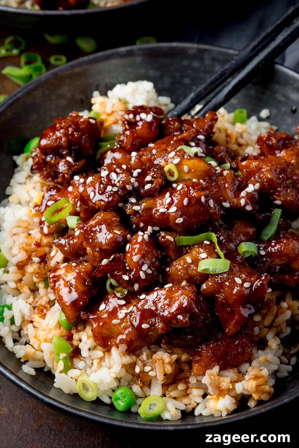 Crispy Air-Fried Sesame Chicken 2 Tall close-up image of air fryer sesame chicken served on a bed of egg fried rice in a black bowl. The sesame chicken is topped with sesame seeds and spring onions. There is a pair of black chopsticks sticking out of the bowl. The bowl is on a dark background.