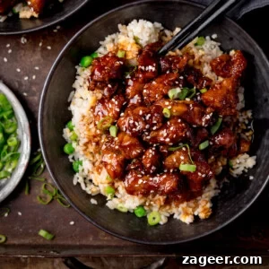 Crispy Air-Fried Sesame Chicken 7 Square image of air fryer sesame chicken served on a bed of egg fried rice in a black bowl. The sesame chicken is topped with sesame seeds and spring onions. There is a pair of black chopsticks sticking out of the bowl. The bowl is on a dark background. There is a small plate with spring onions on the the left of the frame.