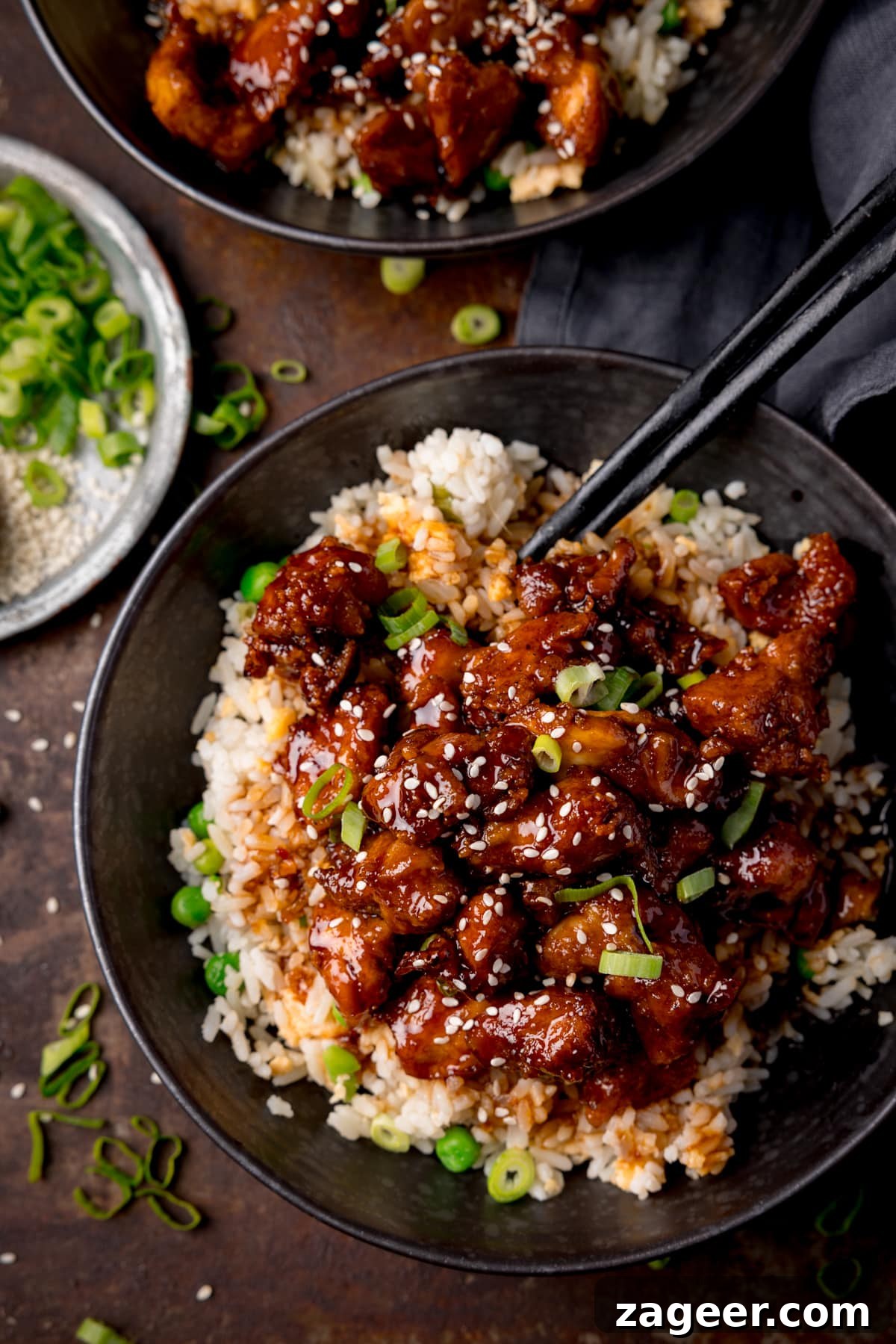 Crispy Air-Fried Sesame Chicken 5 Tall overhead image of air fryer sesame chicken served on a bed of egg fried rice in a black bowl. The sesame chicken is topped with sesame seeds and spring onions. There is a pair of black chopsticks sticking out of the bowl. The bowl is on a dark background. There is a further bowl of sesame chicken at the top of the frame and a small plate with spring onions on the the left of the frame.