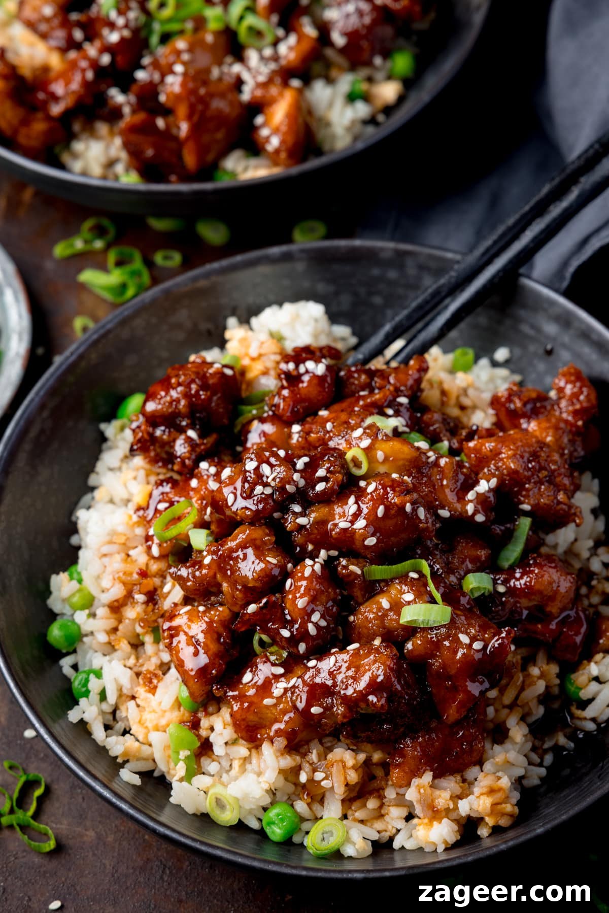 Crispy Air-Fried Sesame Chicken 6 Tall image of air fryer sesame chicken served on a bed of egg fried rice in a black bowl. The sesame chicken is topped with sesame seeds and spring onions. There is a pair of black chopsticks sticking out of the bowl. The bowl is on a dark background. There is a further bowl of sesame chicken at the top of the frame.