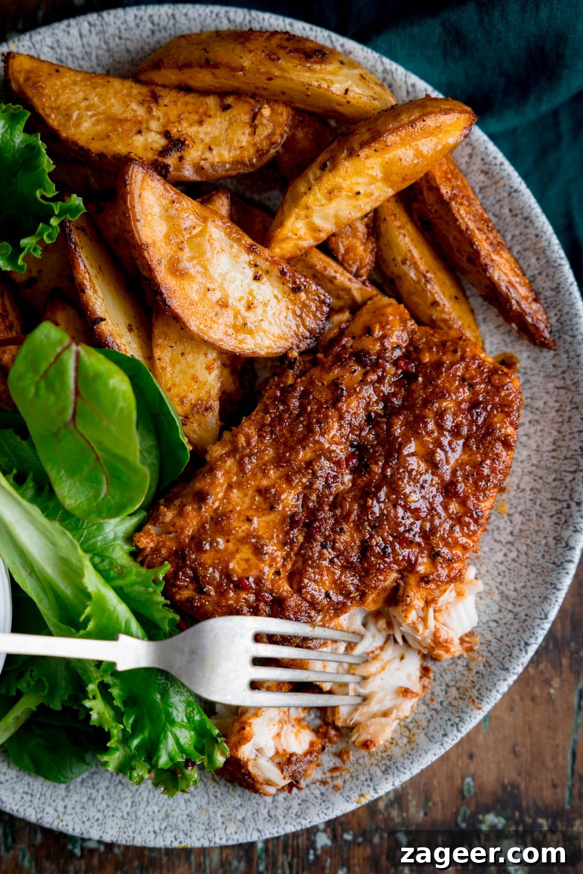 Zesty Cajun Cod and Smoky Roasted Potatoes 5 An inviting overhead view of a grey plate generously laden with Cajun cod, perfectly golden smoky potato wedges, and a side of vibrant green salad leaves. The cod shows beautiful flakiness, with the tip of a silver fork poised to take a bite. The entire setting is on a rich brown background, creating a warm and rustic ambiance.
