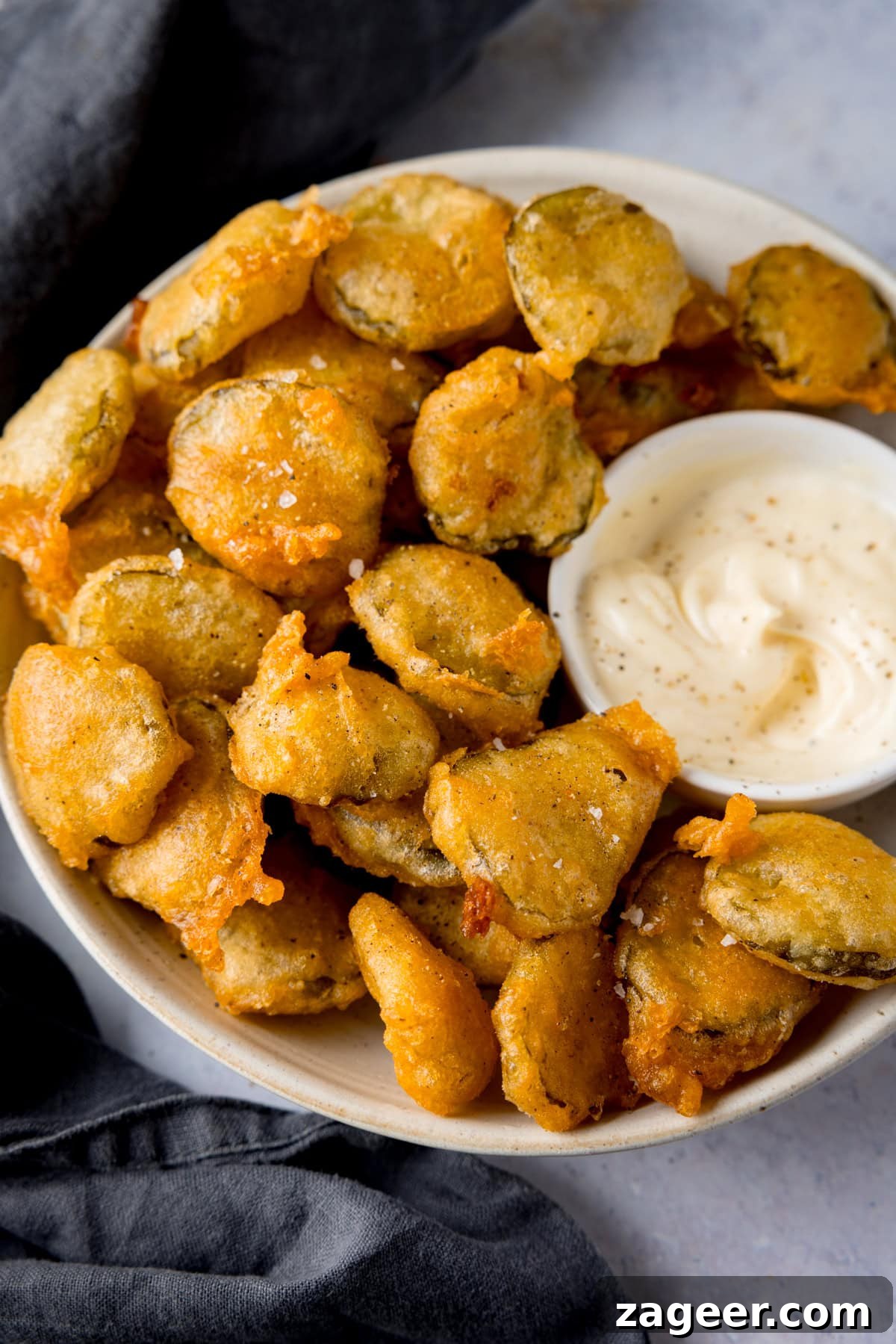 Golden Fried Dill Delights 2 A tall image of many golden-brown fried pickle bites in a white bowl, with a smaller white dish of creamy garlic mayo nestled to the right. A slate grey napkin peeks from under the main bowl, all set against a light grey background.