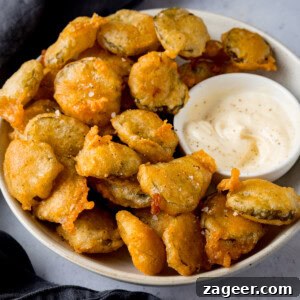 A square image of many fried pickle bites in a white bowl, with a smaller white dish of garlic mayo nestled to the right. A slate grey napkin is visible in the background.