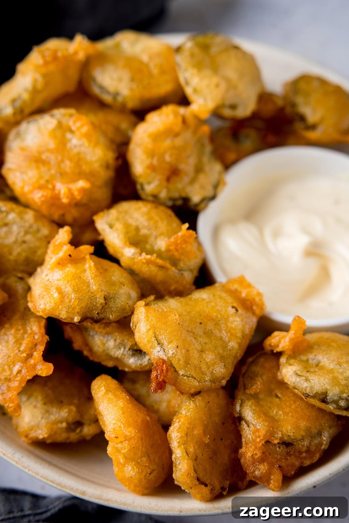 Golden Fried Dill Delights 4 A tall, close-up image of many golden-brown fried pickle bites piled high in a white bowl, with a smaller white dish of creamy garlic mayo nestled into the right side. A slate grey napkin is visible in the background, all resting on a light grey surface.