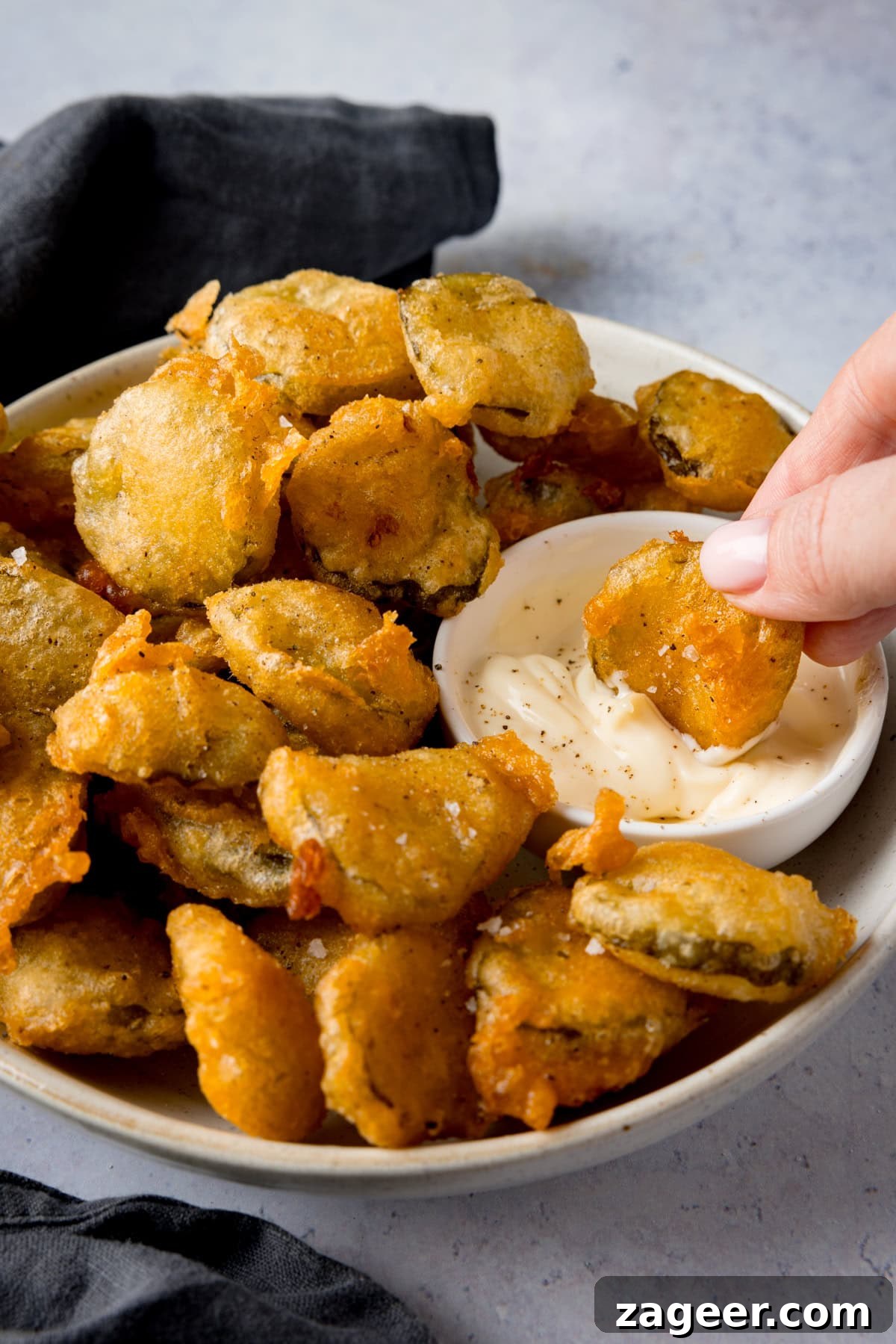 Golden Fried Dill Delights 5 A tall image of many golden-brown fried pickle bites in a white bowl. A smaller white dish of garlic mayo is nestled in the right of the main bowl. A hand is shown dipping a fried pickle into the garlic mayo. A slate grey napkin is visible in the background, all on a light grey surface.