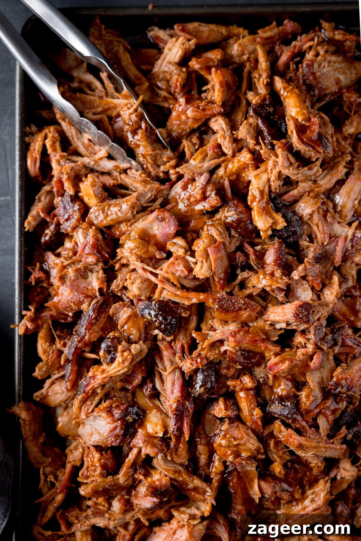 Slow-Cooked BBQ Pork Perfection 2 A tall, overhead image of BBQ Pulled Pork. The pulled pork is in a rectangular black pan in the centre of the image. In the top left of the pan, there is a pair of tongs sticking out of the pork at an angle. This is all set on a grey background.