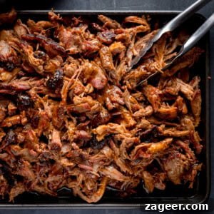 A square, overhead image of BBQ Pulled Pork. The pulled pork is in a rectangular black pan in the centre of the image. In the top right of the pan, there is a pair of tongs sticking out of the pork at an angle. This is all set on a grey background.