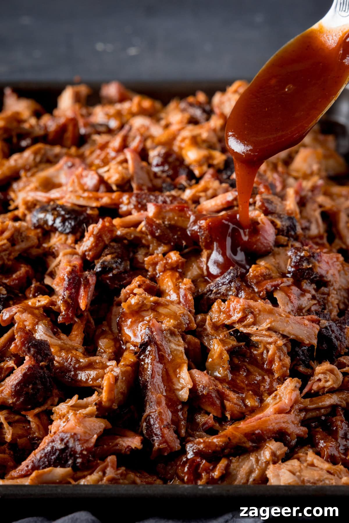 Slow-Cooked BBQ Pork Perfection 5 A tall image of BBQ Pulled Pork in a rectangular black pan. In the top right of the picture, there is a silver spoon with BBQ sauce on it, which is being poured over the Pulled Pork. At the bottom of the image, you can slightly see a navy blue napkin tucked underneath the pan. This is all set on a blue/grey background.