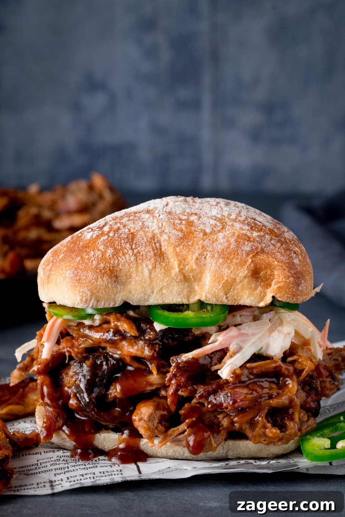 Slow-Cooked BBQ Pork Perfection 6 A tall image of a sandwich, containing the BBQ Pulled pork. The sandwich is in the centre of the image, on top of a piece of white parchment paper. Next to the sandwich is a little bit of pulled pork that has seemingly fallen out of the sandwich, and on the other side, there are two jalapeño slices. On the left side of the background, you can see the rest of the pulled pork in a black skillet. And on the right side of the background, there is a navy blue napkin. This is all set on a grey surface and a blue/grey background.
