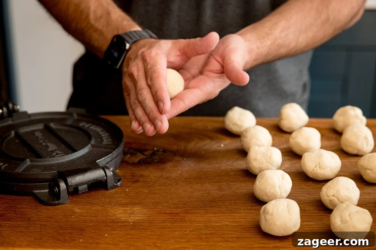 Perfect Homemade Corn Tortillas 5 A cook's hands skillfully rolling a small, smooth ball of corn tortilla dough, positioned next to a black tortilla press and a collection of other dough balls, all on a rustic wooden cutting board, illustrating the precise preparation process.