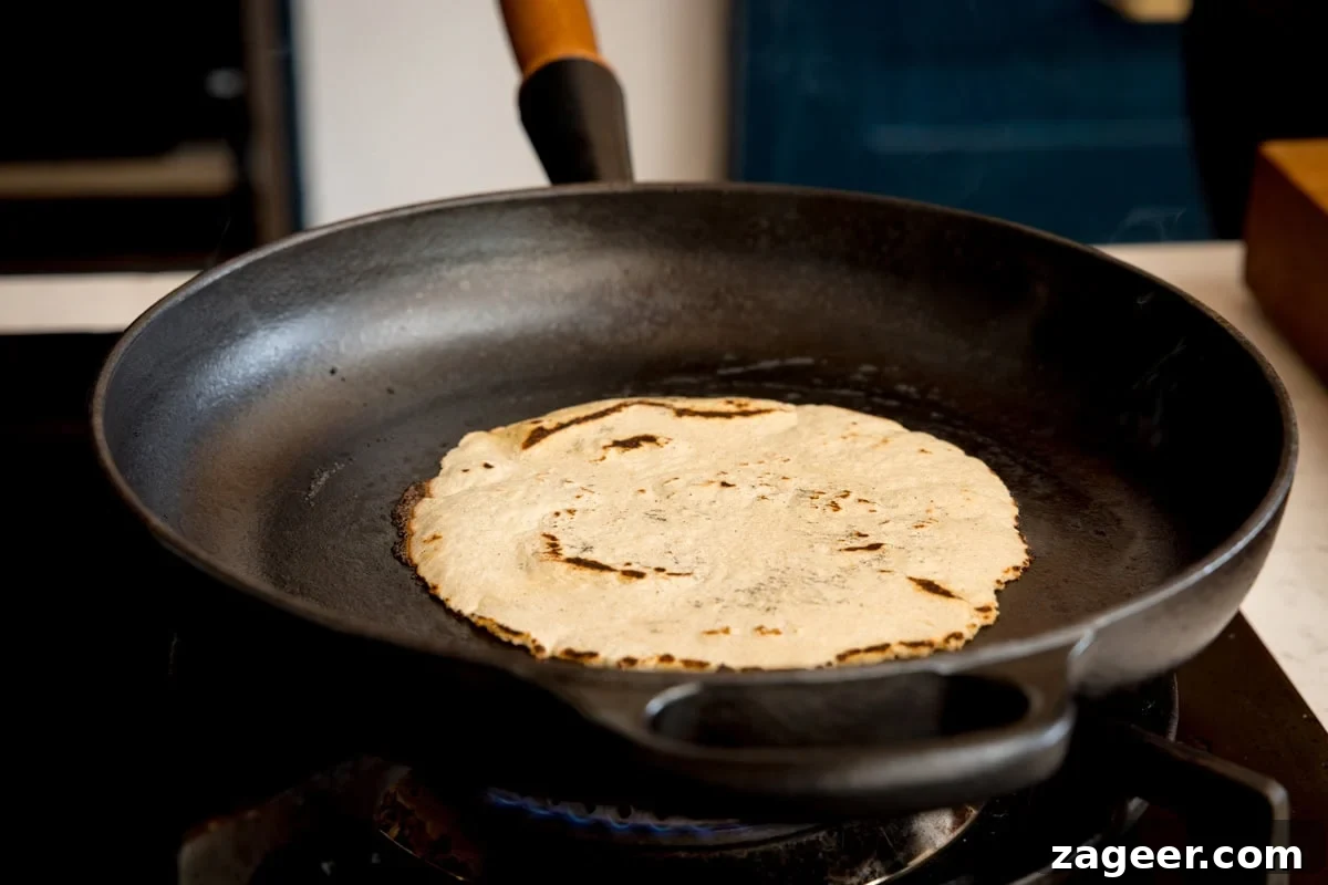 Perfect Homemade Corn Tortillas 7 A single, fresh homemade corn tortilla actively cooking in the center of a black cast iron skillet, positioned on a stovetop burner. The tortilla shows initial charring and hints of puffing, capturing a moment of perfect preparation.