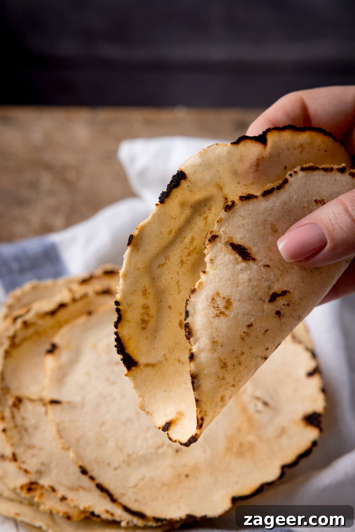 Perfect Homemade Corn Tortillas 8 A close-up square shot of a stack of eight warm, golden corn tortillas. In the foreground, a hand gently folds one of the tortillas in half, showcasing its remarkable pliability. The scene is set on a rustic wooden surface with a blue and white striped napkin, enhancing the homemade feel.