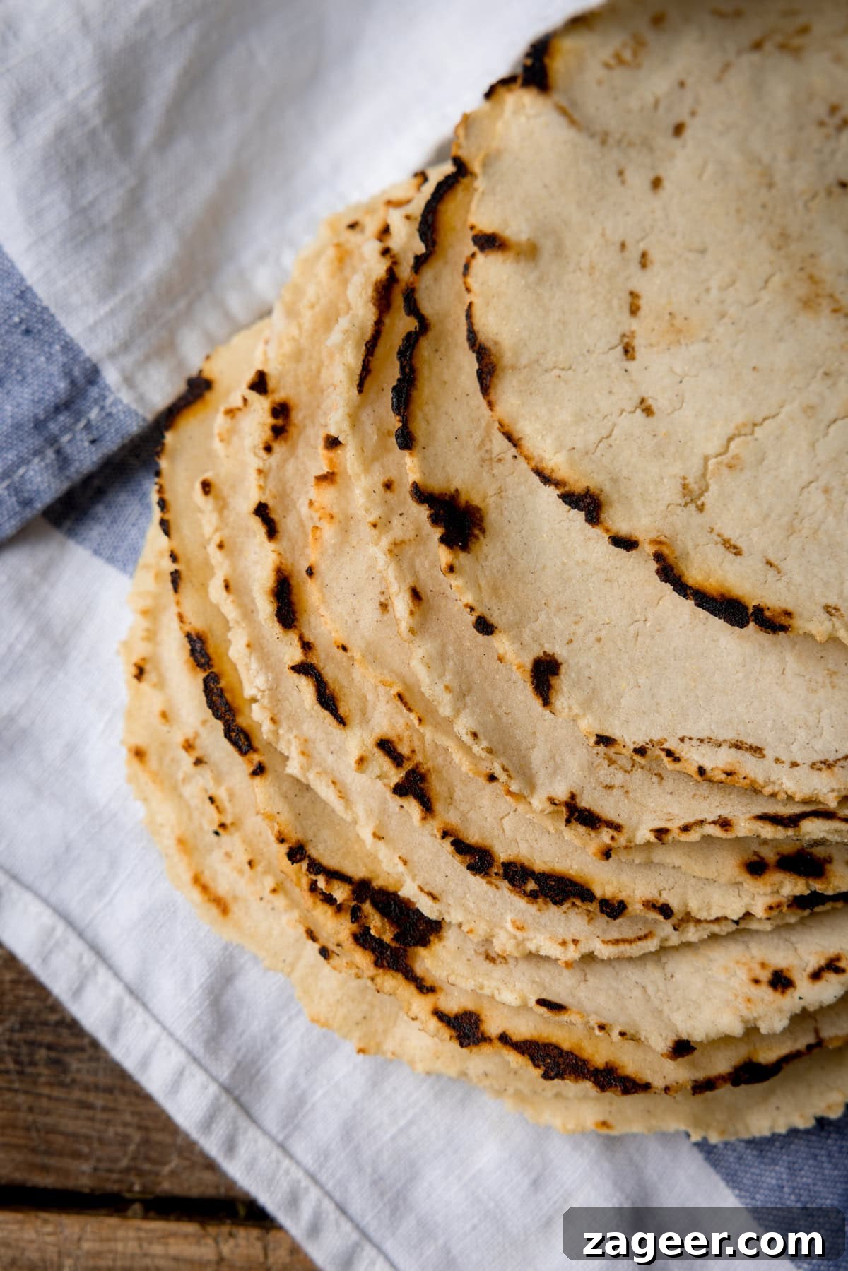 Perfect Homemade Corn Tortillas 9 A tall, inviting overhead shot of a perfectly arranged stack of nine golden-yellow homemade corn tortillas, elegantly presented on a crisp blue and white striped napkin, all set against a warm, rustic wooden surface, highlighting their artisanal charm.