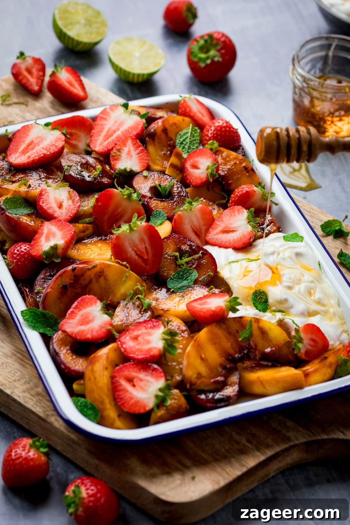 Sizzling Fruit Delight 2 A tall, inviting shot of vibrant griddled fruit salad arranged in a white tray with a blue lining, centered on a wooden cutting board. A generous dollop of creamy Greek yogurt sits gracefully on the bottom right. Fresh, ruby-red strawberries are scattered artfully around the board. In the top right, a small glass bowl gleams with golden honey, beside a honey stirrer dripping with its sweet contents onto the fruit. A few bright lime wedges add a final touch of freshness to this delightful summer spread, all set against a soft, light grey background.