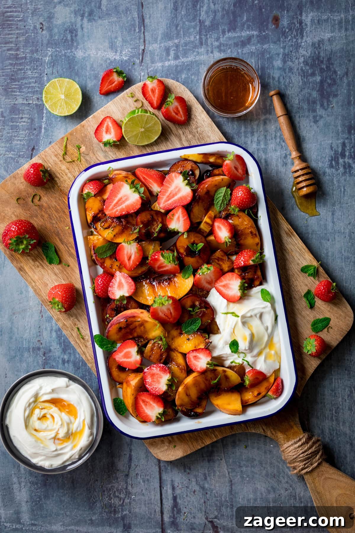Sizzling Fruit Delight 3 A tall, overhead shot of a vibrant griddled fruit salad nestled in a white tray with a blue lining, resting at an angle on a rustic wooden cutting board. Scattered around the board are fresh strawberries, adding a pop of color. To the left, a small black bowl holds more creamy Greek yogurt, elegantly topped with a swirl of honey. In the top right corner, a glass bowl shimmers with golden honey, accompanied by a honey stirrer coated in its sticky sweetness. A few bright lime wedges are also visible, one on the wooden board and another beside it. This entire arrangement is set against a subtle, light grey background.