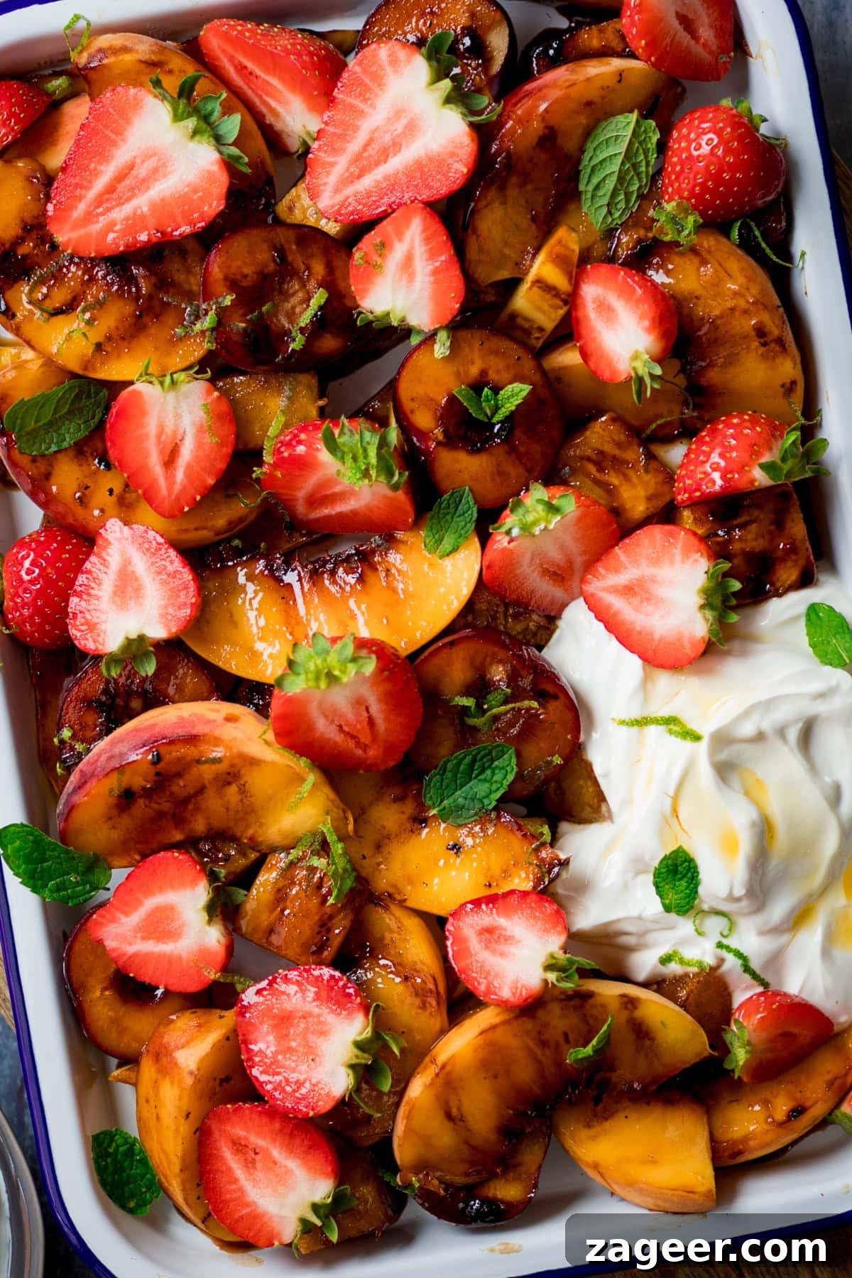 Sizzling Fruit Delight 4 A tall, close-up image showcasing a vibrant griddled fruit salad within a white tray featuring a blue lining. In the bottom right corner of the tray, a perfectly formed dollop of creamy Greek yogurt adds a refreshing touch. Small, delicate mint leaves are artfully scattered atop the fruit, enhancing both its visual appeal and aromatic freshness. The image focuses intensely on the tray of fruit, highlighting the caramelized textures and bright colors.