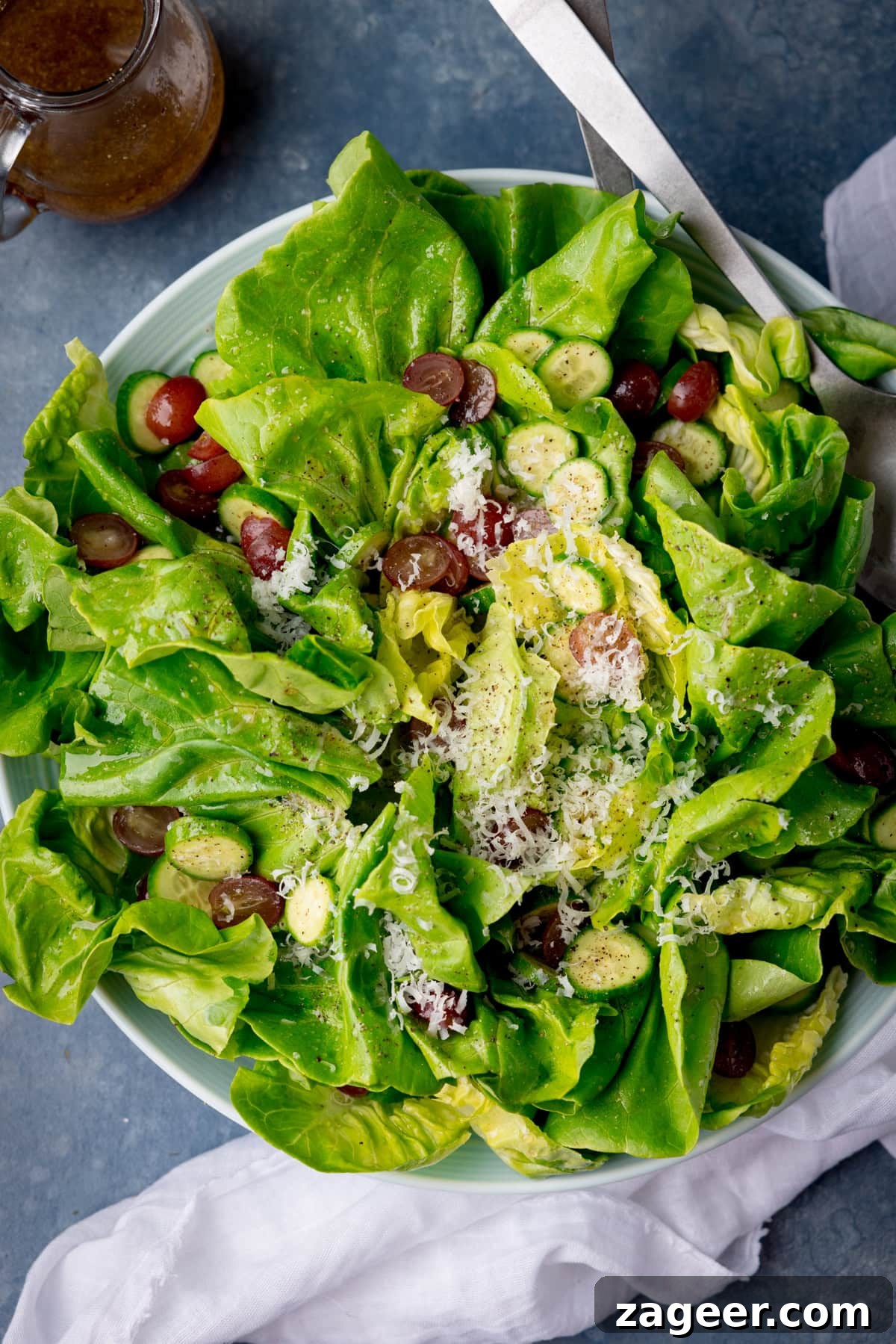 Velvety Butter Lettuce Salad 2 A tall, overhead shot of Butter Lettuce Salad in a large white bowl. The bowl with the salad is placed in the centre of the image, in the top right corner of the bowl there are two silver metal salad salad servers sticking out of the dish. In the top left corner of the image, you can see a clear glass jug with the rest of the salad dressing in it. Just tucked below the bottom right-hand side of the bowl, there is a white napkin. This is all set on a light blue/grey background.
