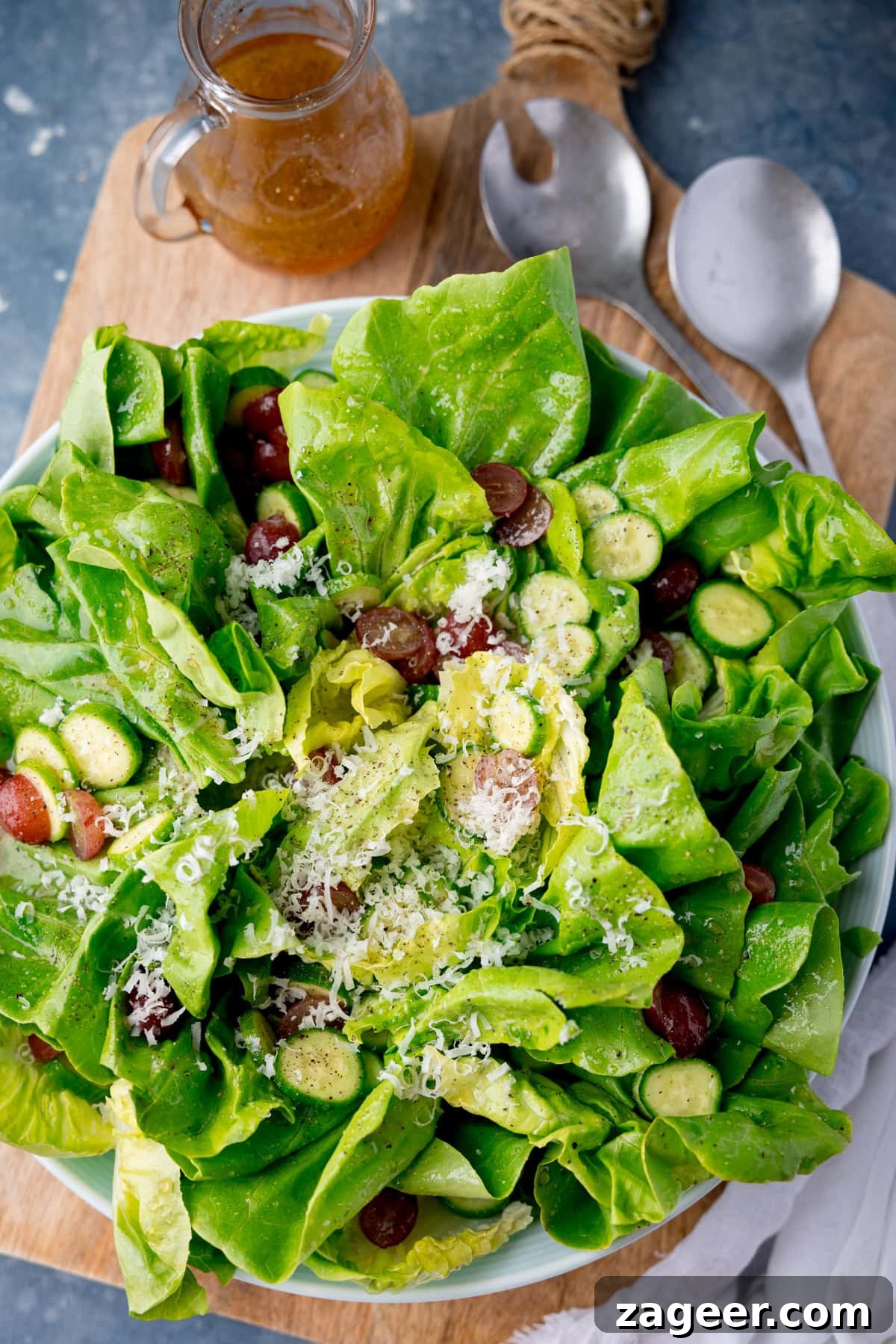 Velvety Butter Lettuce Salad 3 A tall, overhead shot of Butter Lettuce Salad in a large white bowl. The bowl with the salad is placed in the centre of the image on top of a large wooden cutting board, on the top right corner of the cutting board, beside the bowl, there are two silver metal salad salad servers sticking out of the dish. In the top left corner of the image, you can see a clear glass jug with the rest of the salad dressing in it. Just tucked below the bottom right-hand side of the bowl, there is a white napkin. This is all set on a light blue/grey background.
