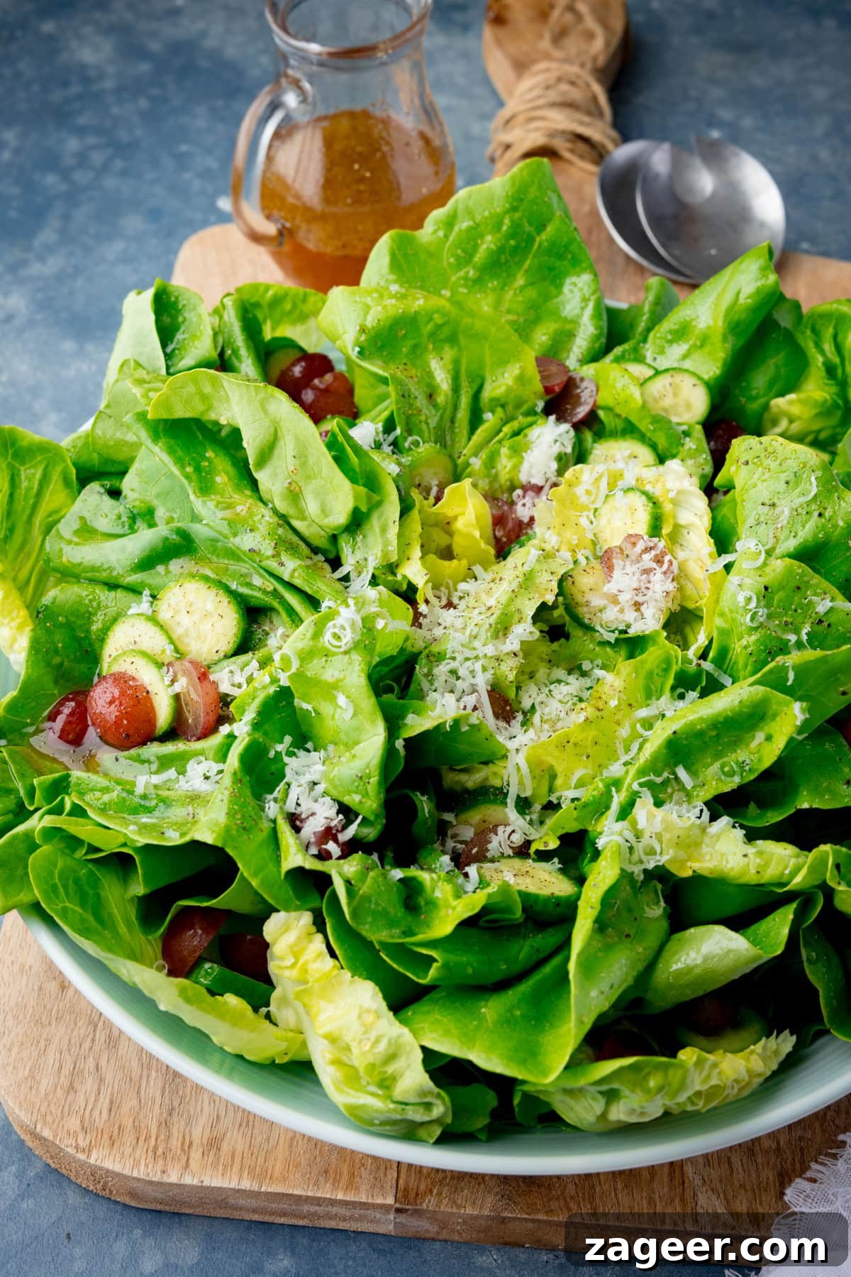 Velvety Butter Lettuce Salad 4 A tall shot of Butter Lettuce Salad in a large white bowl. The bowl with the salad is placed in the centre of the image on top of a large wooden cutting board, on the top right corner of the cutting board, beside the bowl, there are two silver metal salad salad servers sticking out of the dish. In the top left corner of the image, you can see a clear glass jug with the rest of the salad dressing in it. In the bottom left corner of the image, you can ever so slightly see a white napkin. This is all set on a light blue/grey background.