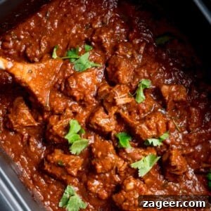 Hearty Slow Cooker Pork and Beef Chili 6 Square close-up overhead image of slow cooker pork and beef chilli in a slow cooker with a wooden spoon sticking out. There is a little sprinkle fresh coriander on top.
