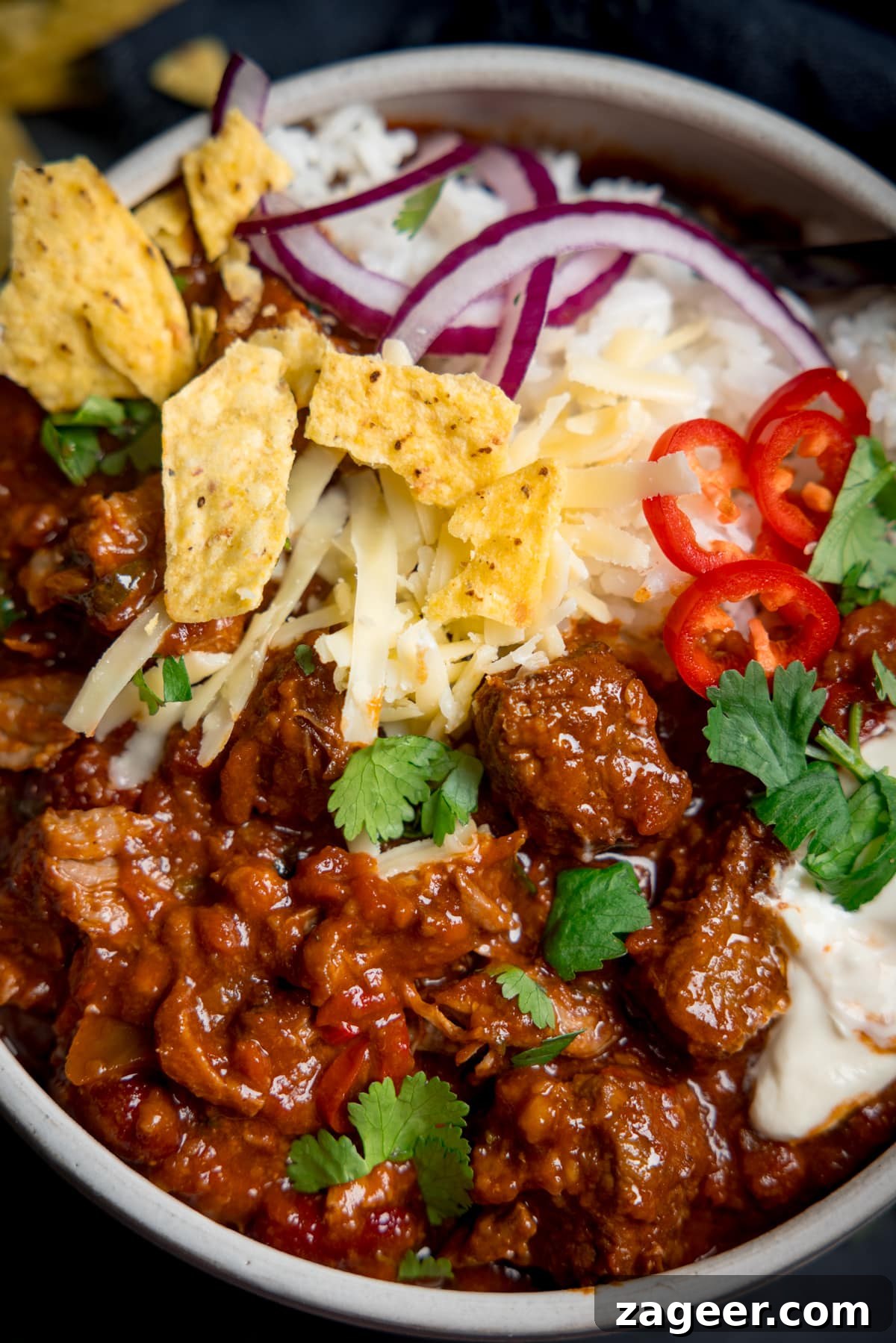 Hearty Slow Cooker Pork and Beef Chili 4 Overhead of pork and beef chilli in bowl with rice, sour cream, sliced red chillies, grated cheese, sliced red onion, fresh coriander and crumbled tortilla chips on top.