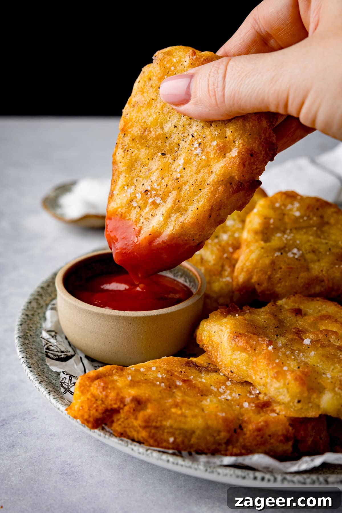 Dipping a Homemade Hash Brown into Ketchup A tall stack of six golden Air Fryer Hash Browns on a plate with newspaper parchment. A hand holds one hash brown, dipping it into a small pot of ketchup. A blue-striped napkin and a dish of sea salt are in the background against a white setting.