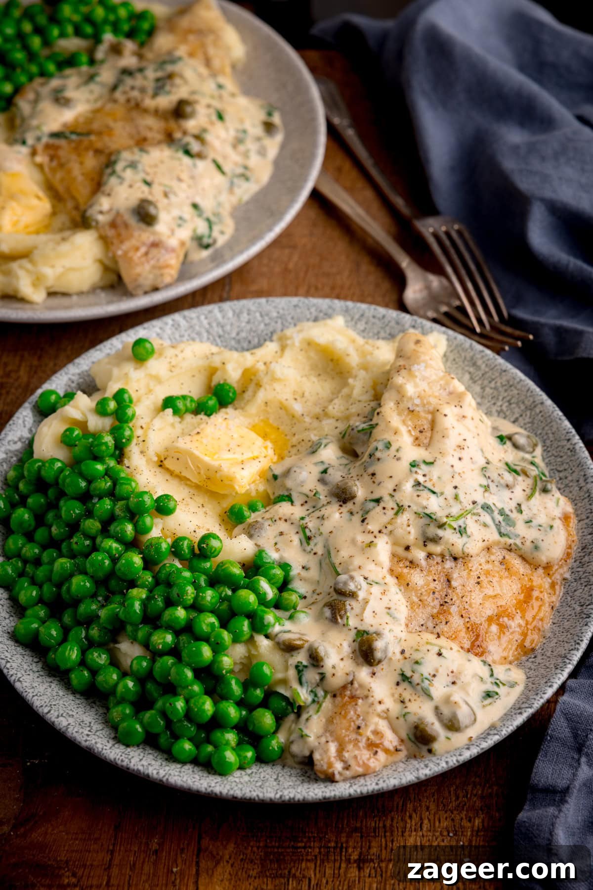 Zesty Lemon Butter Sea Bream 2 A plate of lemon butter sea bream in creamy sauce, mashed potatoes, and peas on a wooden table. Nearby is a cloth napkin, a second plate of the sea bream and 2 forks.