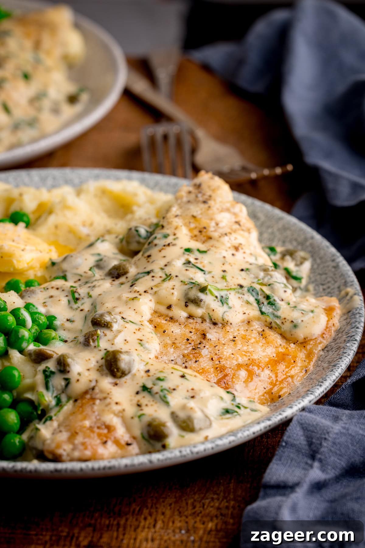 Zesty Lemon Butter Sea Bream 5 A side-on image of a plate of lemon butter sea bream in a creamy sauce with capers served with mashed potatoes and peas. An extra plate and cutlery are visible in the background on a wooden surface.