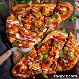 Flatbread pizzas topped with grilled chicken tikka, a rich tomato sauce, fresh coriander, crunchy sliced almonds, and a delicate drizzle of creamy white sauce. This appealing chicken tikka naan pizza is perfectly presented on a wooden board alongside a pizza cutter.