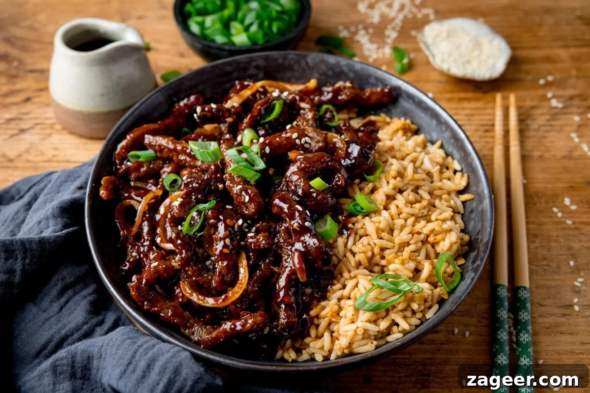 Crispy Air-Fried Chili Beef 12 A wide image of a black bowl filled with air fryer crispy chilli beef with onions, sesame seeds, and sliced spring onions, served with fried rice. The bowl is on a wooden surface next to a blue/grey napkin, a pair of chopsticks, a small jug of soy sauce, a small bowl of chopped spring onions and a small dish of sesame seeds.