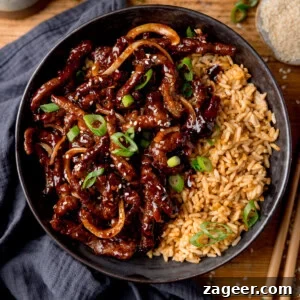 A black bowl filled with air fryer crispy chilli beef with onions, sesame seeds, and sliced spring onions, served with fried rice. The bowl is on a wooden surface next to a blue/grey napkin, a pair of chopsticks and a small dish of sesame seeds.
