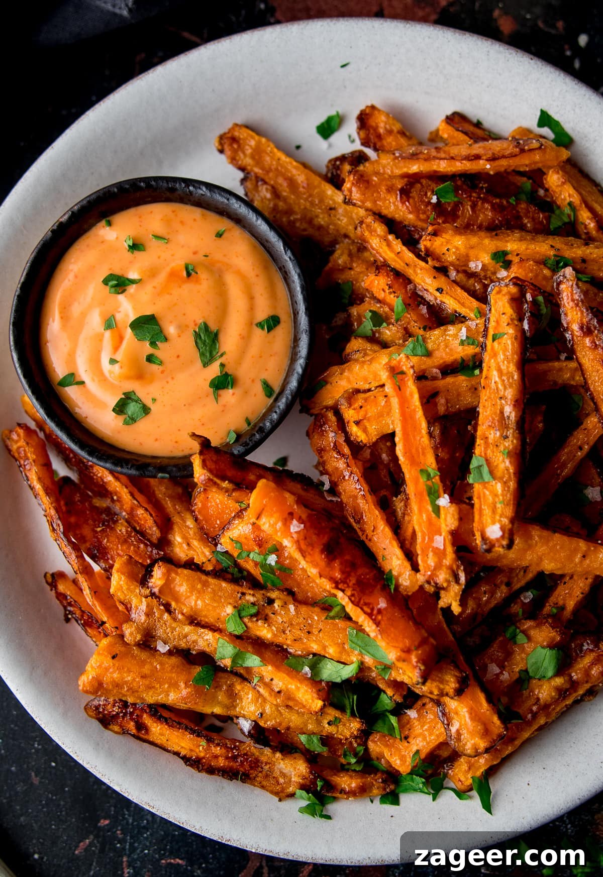 Golden Parmesan Carrot Fries 2 Close up overhead shot of golden-brown baked parmesan carrot fries on a white plate with a small bowl of vibrant red sriracha mayo dipping sauce. Garnished with fresh green herbs.