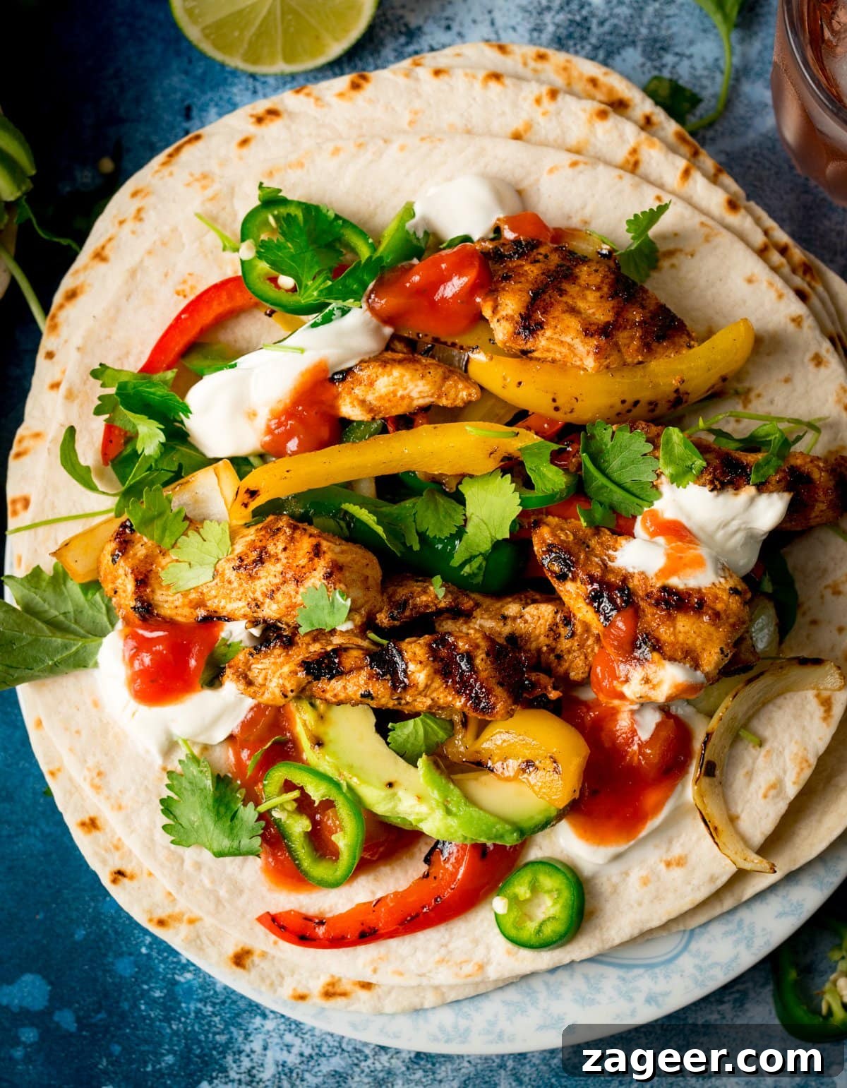 Flavorful Chicken Fajitas Made Easy 4 Two perfectly assembled flour tortillas, generously topped with grilled fajita chicken, vibrant peppers, caramelized onions, creamy avocado, fiery jalapeño slices, fresh coriander, tangy salsa, and a dollop of cool soured cream, presented on a rich blue background.