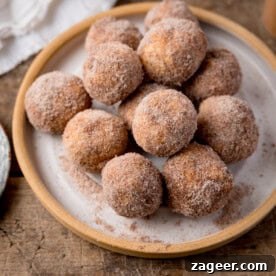 A beautifully arranged stack of perfectly golden air fryer doughnut holes, generously coated in shimmering cinnamon sugar, presented invitingly on a pristine white plate with a subtle light brown rim. This delightful stack rests on a warm wooden surface, suggesting a cozy and delicious homemade treat.