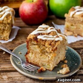 A square image beautifully showcasing a generous slice of decadent apple streusel cake, adorned with a luscious, artistic drizzle of sweet vanilla icing. The distinct layers of soft cake, spiced apple, and crunchy streusel are visible, promising a delightful texture and rich flavour experience.