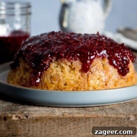 A classic, golden slow cooker steamed jam sponge cake, fresh from the cooker, resting invitingly on a grey plate. The plate is positioned on a rustic wooden chopping board, highlighting the homely charm of this comforting dessert.