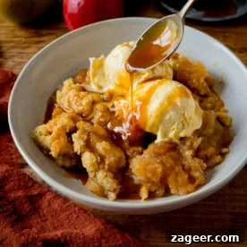A white bowl filled with golden apple crumble, beautifully topped with two scoops of creamy vanilla ice cream. A stream of rich caramel sauce is being drizzled over with a spoon. The inviting bowl rests on a rustic wooden table, accompanied by a dark orange napkin and a couple of fresh, whole apples in the background.