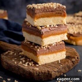 Three perfectly cut, decadent pieces of salted caramel millionaire's shortbread are artfully piled on a rustic wooden board, set against a dark, moody background. Each piece showcases distinct layers of shortbread, caramel, and chocolate, hinting at rich indulgence.