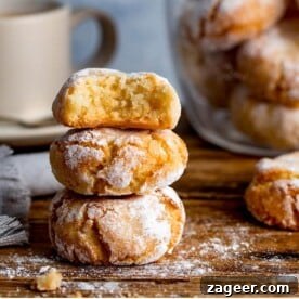 A tempting pile of Italian Amaretti cookies, beautifully crinkled and dusted with icing sugar, artfully arranged on a rustic wooden board. A blurred cup in the background subtly suggests a delightful coffee pairing, inviting a moment of sweet indulgence.