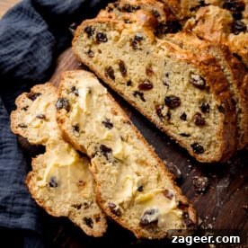 Several inviting slices of fluffy fruit soda bread, generously spread with creamy butter, gracefully rest on a rustic wooden surface. A dark, textured cloth is draped nearby, adding to the cozy and inviting aesthetic of a wholesome breakfast or snack.
