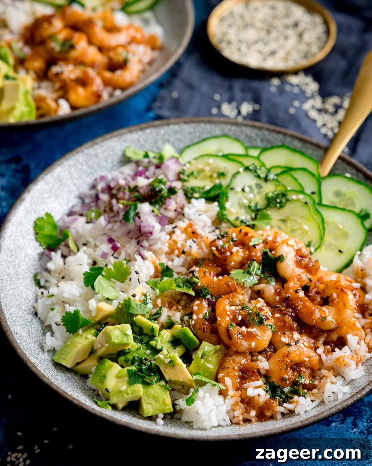 Garlic Honey Shrimp Rice Bowls 5 A bowl with white rice, cooked prawns in honey garlic sauce, diced avocado, chopped red onion, cucumber slices, coriander, and sesame seeds on top.