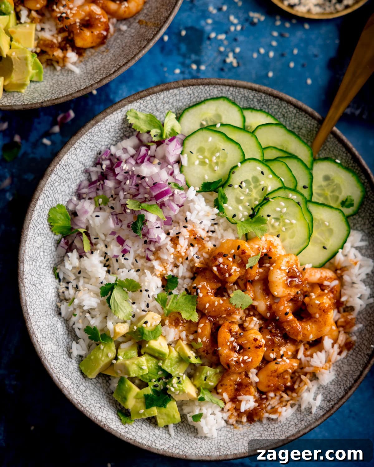 Garlic Honey Shrimp Rice Bowls 6 A bowl with white rice, cooked honey garlic prawns in sauce, sliced cucumber, diced red onion, avocado chunks, coriander, and sesame seeds on a blue background.