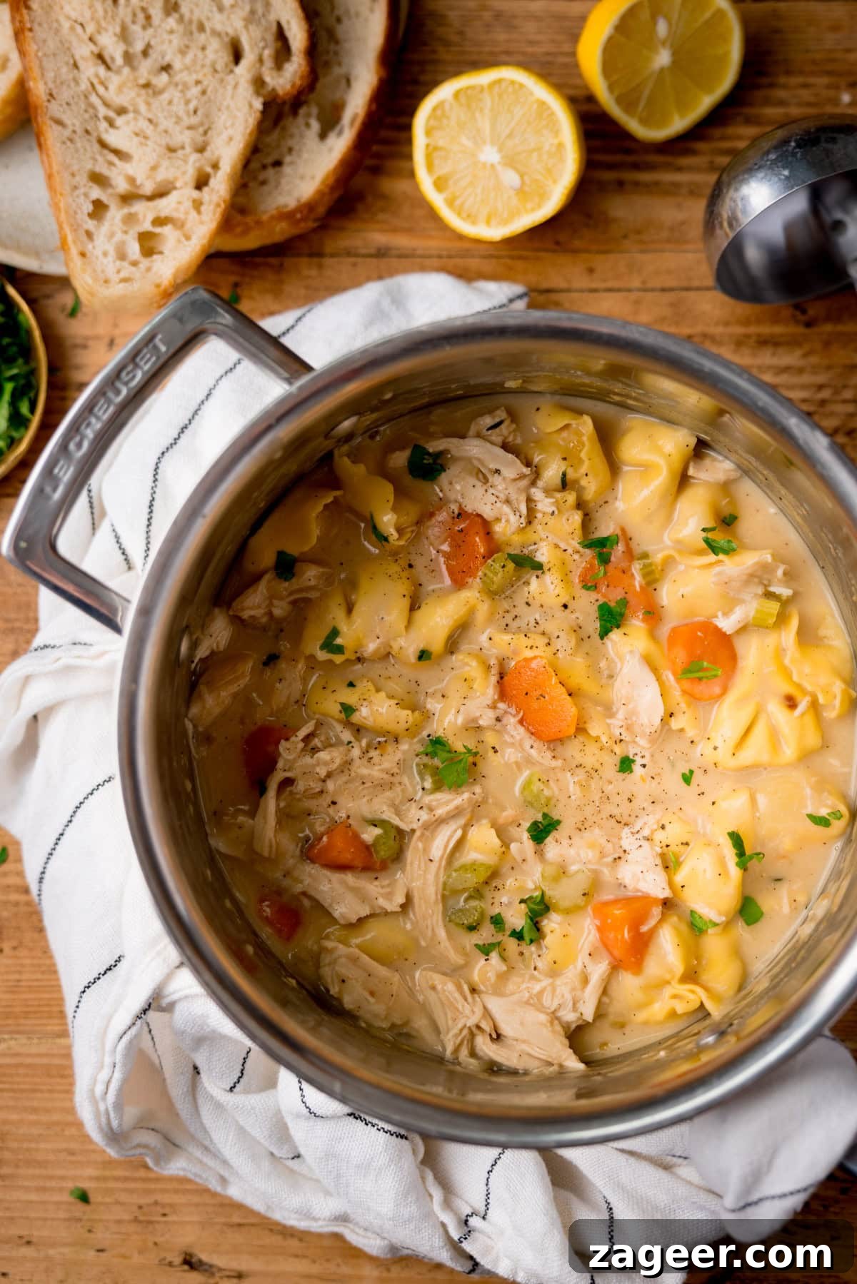 Velvet Chicken Tortellini Comfort 5 A large pot of creamy chicken and tortellini soup, brimming with carrots and fresh parsley, rests on a wooden table. Slices of artisanal bread and vibrant lemon halves are arranged nearby, highlighting a comforting and inviting meal setting.