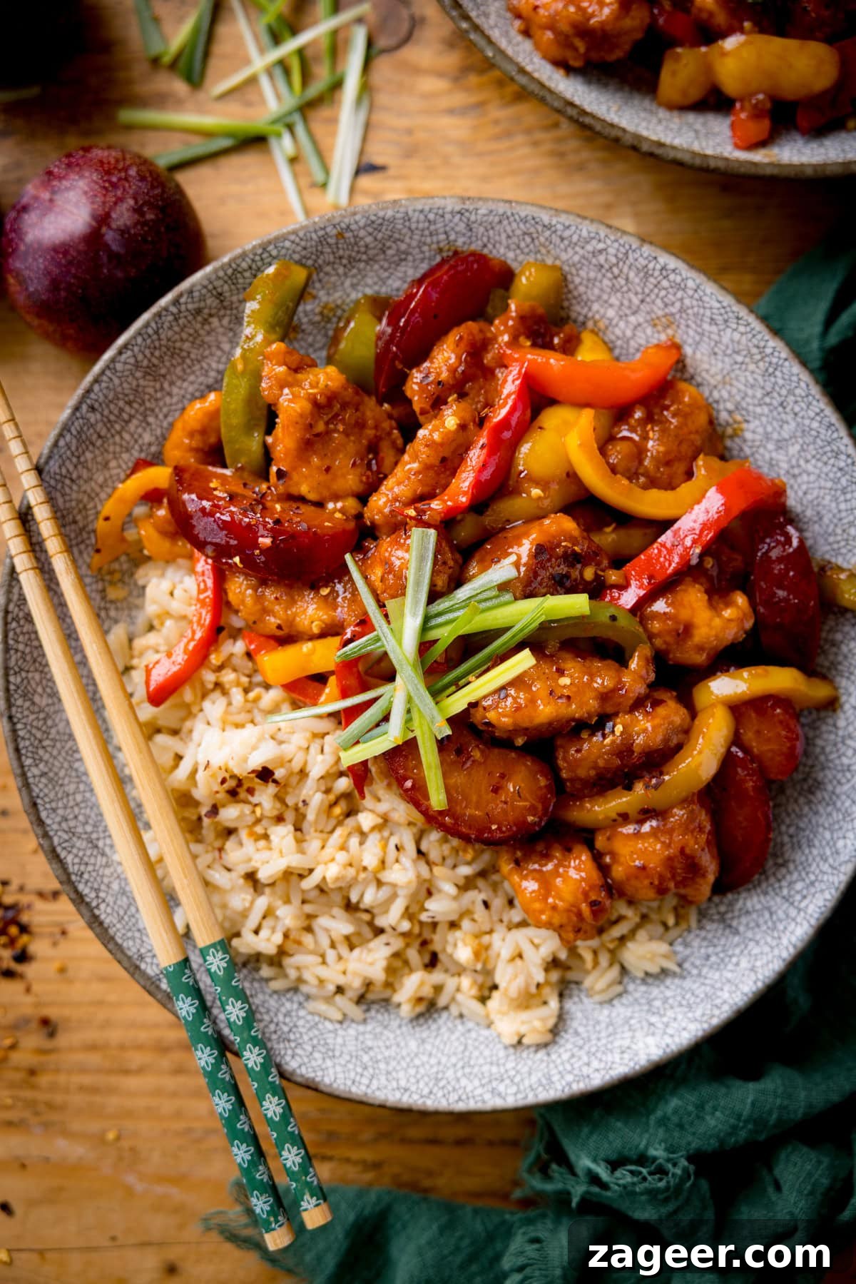 Imperial Plum Chicken Stir-Fry 6 Chinese plum chicken stir fry and rice in a shallow bowl with strips of spring onion on top. There is a set of chopsticks on the side of the bowl. The bowl is on a wooden table next to a green napkin.