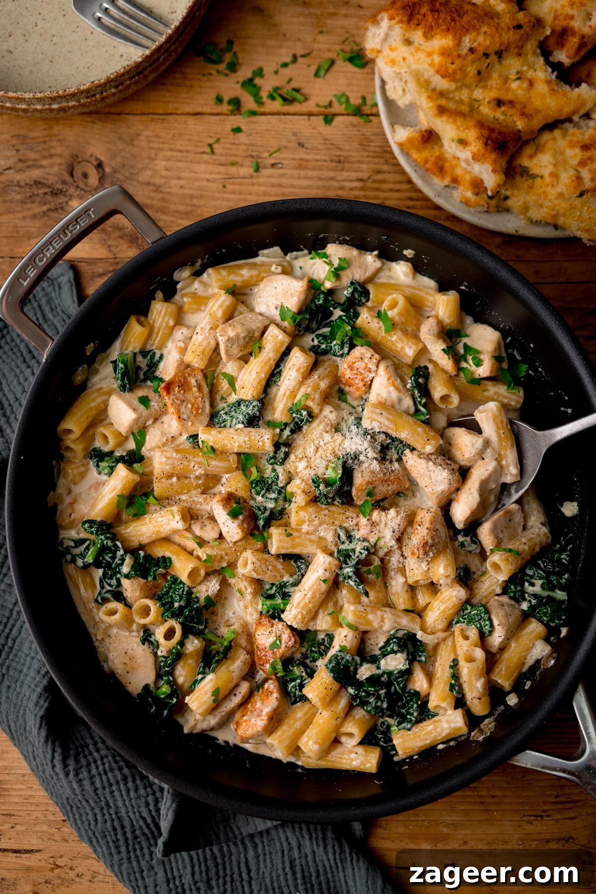 One-Pot Creamy Chicken Rigatoni Alfredo 5 A frying pan filled with creamy rigatoni alfredo pasta, chicken pieces, and kale, topped with grated cheese and parsley. Garlic bread is on a plate nearby.